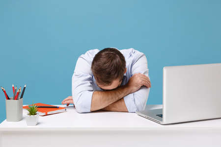 Tired young man in light shirt sit and work at desk with pc laptop isolated on pastel blue...