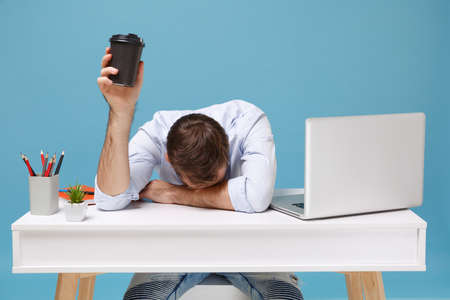 Tired young man in shirt sit and work at desk with pc laptop isolated on blue background. Achievement business career concept. Mock up copy space. Laid his head down on desk hold cup of coffee or tea.の写真素材