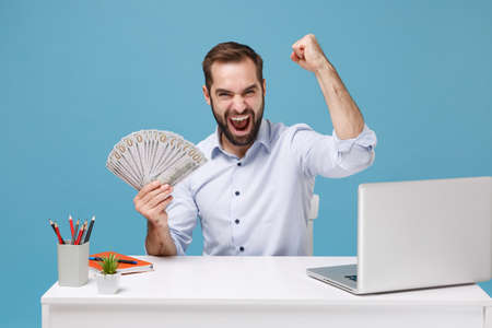 Happy man in shirt work at desk with pc laptop isolated on blue background. Achievement business career concept. Mock up copy space. Holding fan of cash money in dollar banknotes doing winner gesture.の写真素材