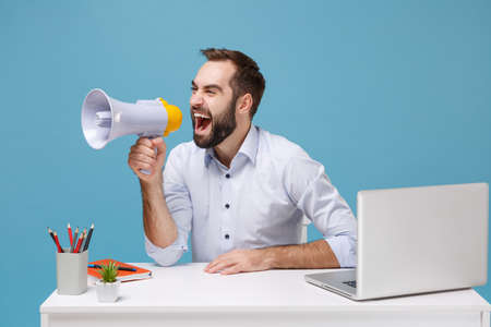 Crazy young bearded man in light shirt sit work at desk with pc laptop isolated on pastel blue background. Achievement business career concept. Mock up copy space. Scream in megaphone, looking aside.の写真素材