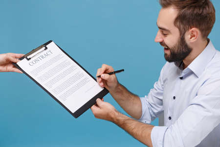 Cheerful young bearded man in light shirt work isolated on pastel blue background in studio. Achievement business career concept. Mock up copy space. Signing clipboard with paper document contract.の写真素材
