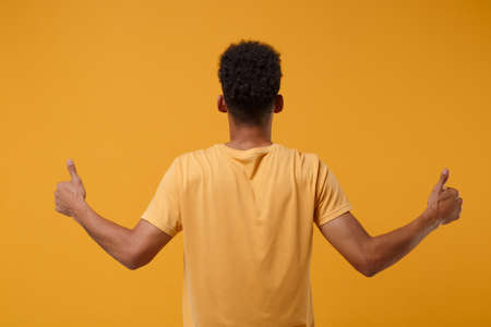 Back rear view of young african american guy in casual t-shirt posing isolated on yellow orange wall...
