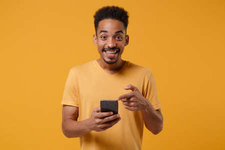 Funny young african american guy in casual t-shirt posing isolated on yellow orange background, studio portrait. People lifestyle concept. Mock up copy space. Pointing index finger on mobile phone.の写真素材