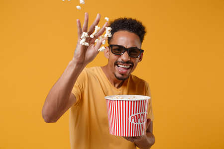 Laughing young african american guy in 3d glasses posing isolated on yellow orange background. People lifestyle concept. Mock up copy space. Watching movie film, holding bucket, throwing popcorn.の写真素材