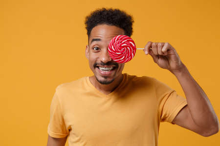 Smiling young african american guy in casual t-shirt posing isolated on yellow orange background studio portrait. People emotions lifestyle concept. Mock up copy space. Cover eye with round lollipop.の写真素材