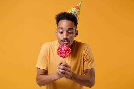 Funny young african american guy in casual t-shirt, birthday hat posing isolated on yellow orange...