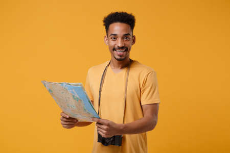 Smiling young african american guy in casual t-shirt with retro vintage photo camera posing isolated on yellow orange background. People lifestyle concept. Mock up copy space. Hold in hands city map.の写真素材