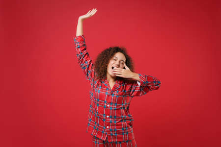 Relaxed african american girl in pajamas homewear resting at home isolated on red background. Relax good mood lifestyle concept. Keeping eyes closed, rising hand, yawning, covering mouth with palm.の写真素材