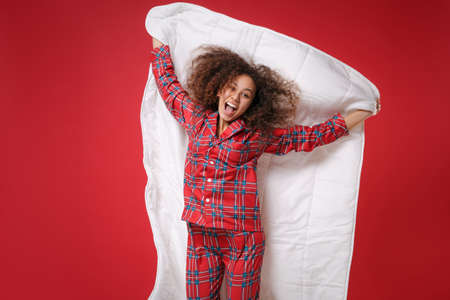 Funny young african american girl in pajamas homewear posing while resting at home isolated on red background. Relax good mood lifestyle concept. Mock up copy space. Jumping with blanket, having fun.の写真素材