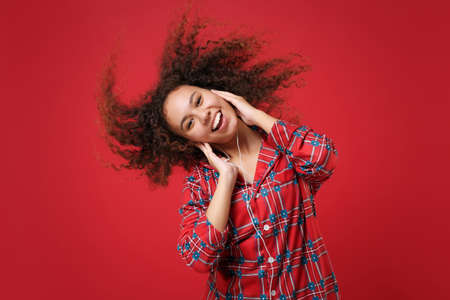 Funny young african american girl in pajamas homewear posing resting at home isolated on red background. Relax good mood lifestyle concept. Mock up copy space. Listen music with earphones, dancing.の写真素材