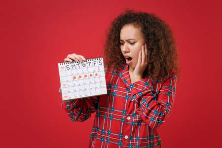 Shocked african american girl in pajamas homewear rest at home isolated on red background. Relax good mood lifestyle concept Hold female periods calendar checking menstruation days put hand on cheek.の写真素材