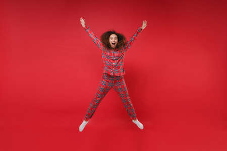 Excited young african american girl in pajamas homewear birthday hat resting at home isolated on red background. Relax good mood lifestyle concept. Mock up copy space. Jumping spreading hands, legs.の写真素材