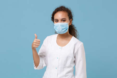 African american doctor woman isolated on blue background. Female doctor in white medical gown, sterile face mask showing thumb up. Healthcare personnel health medicine concept. Mock up copy space.の写真素材