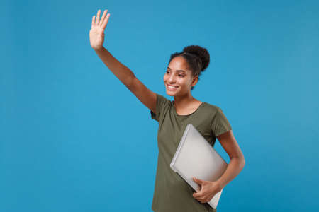 Funny african american woman girl in casual t-shirt isolated on blue background. People lifestyle concept. Mock up copy space. Hold laptop pc computer waving and greeting with hand as notices someone.の写真素材
