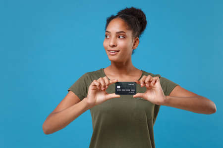 Pretty young african american woman girl in casual t-shirt posing isolated on bright blue background studio portrait. People lifestyle concept. Mock up copy space. Hold credit bank card looking aside.の写真素材