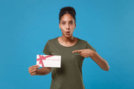 Shocked young african american woman girl in casual t-shirt posing isolated on blue background studio portrait. People lifestyle concept. Mock up copy space. Pointing index finger on gift certificate.の写真素材