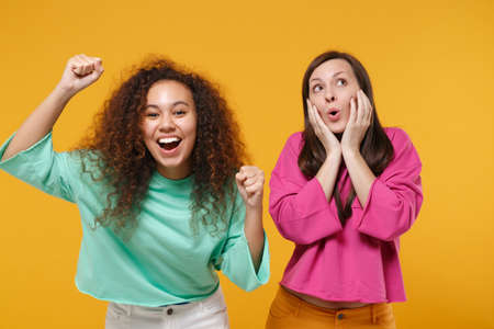 Two funny women friends european african american girls in pink green clothes posing isolated on yellow background. People lifestyle concept. Mock up copy space. Clenching fists put hands on cheeks.の写真素材