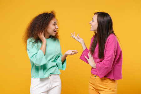 Two amazed women friends european and african american girls in pink green clothes posing isolated on yellow background. People lifestyle concept. Mock up copy space. Looking at each other speaking.の写真素材