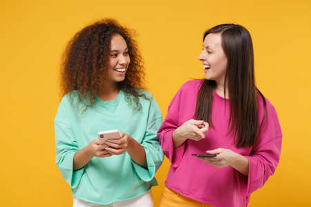Two laughing women friends european african american girls in pink green clothes posing isolated on yellow background. People lifestyle concept. Mock up copy space. Using mobile phone, typing message.の写真素材