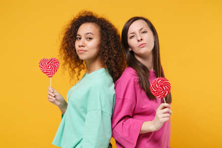 Two beautiful women friends european and african american girls in pink green clothes posing isolated on yellow background. People lifestyle concept. Mock up copy space. Hold round heart lollipops.の写真素材