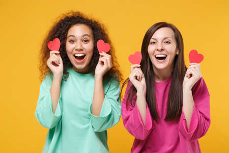 Two excited women friends european and african american girls in pink green clothes posing isolated on yellow wall background. People lifestyle concept. Mock up copy space. Holding little red hearts.の写真素材