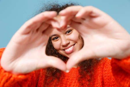 Smiling young african american girl in casual orange knitted clothes isolated on pastel blue background. People lifestyle concept. Mock up copy space. Showing shape heart with hands, heart-shape sign.の写真素材