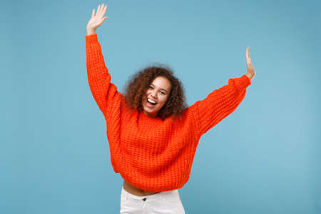 Cheerful young african american girl in casual orange knitted clothes isolated on pastel blue wall background studio portrait. People lifestyle concept. Mock up copy space. Rising spreading hands.の写真素材