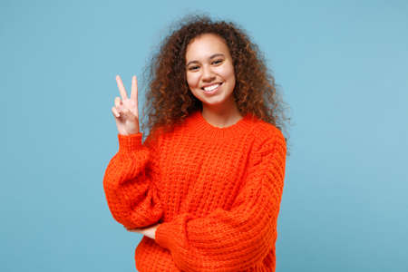 Smiling young african american girl in casual orange knitted clothes isolated on pastel blue background studio portrait. People emotions lifestyle concept. Mock up copy space. Showing victory sign.の写真素材