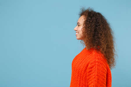 Side view of smiling young african american girl in casual orange knitted clothes isolated on pastel...