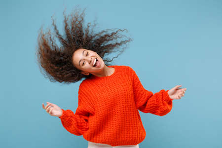 Cheerful young african american girl in casual orange knitted clothes isolated on pastel blue background studio portrait. People lifestyle concept. Mock up copy space. Having fun with fluttering hair.の写真素材