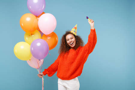 Laughing young african american girl in casual orange knitted clothes, birthday hat isolated on pastel blue wall background. Holiday party concept. Celebrating, holding colorful air balloons, pipe.の写真素材