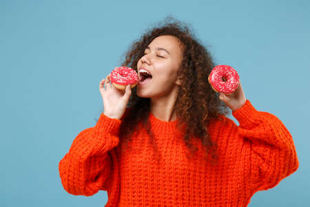 Pretty young african american girl in casual orange knitted clothes isolated on pastel blue background studio portrait. People emotions lifestyle concept. Mock up copy space. Hold biting pink donuts.の写真素材