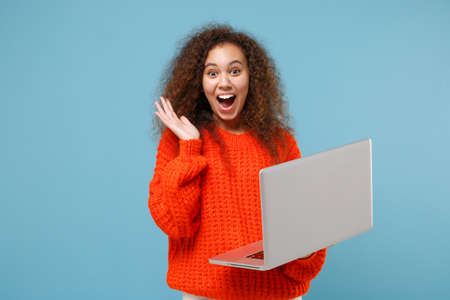 Shocked young african american girl in casual orange knitted clothes isolated on pastel blue background studio portrait. People lifestyle concept. Mock up copy space. Working on laptop pc computer.の写真素材
