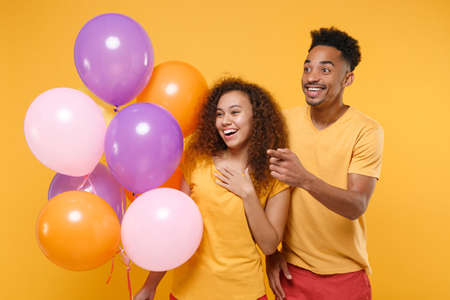 Funny young friends couple african american guy girl in casual clothes isolated on yellow background. Birthday holiday party concept. Celebrating hold colorful air balloons point index finger aside.の写真素材