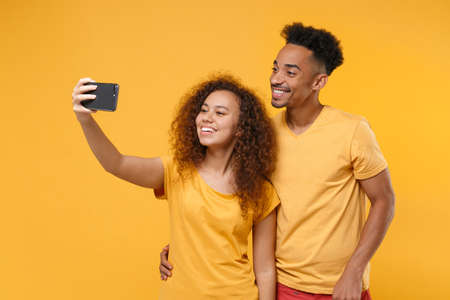 Smiling young friends couple african american guy girl in casual clothes isolated on yellow orange background. People emotions lifestyle concept. Mock up copy space. Doing selfie shot on mobile phone.の写真素材