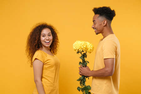 Side view of excited young friends couple african american guy girl in casual clothes isolated on yellow orange wall background. People lifestyle concept. Mock up copy space. Hold bouquet of flowers.の写真素材