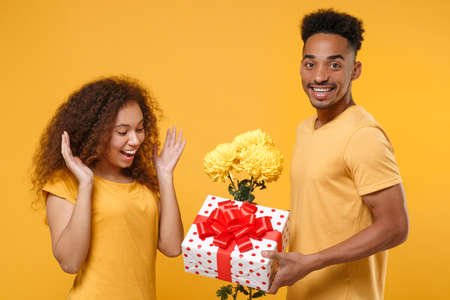 Side view of excited friends couple african american guy girl in casual clothes isolated on yellow background. People lifestyle concept. Hold bouquet of flowers, red present box with gift ribbon bow.の写真素材