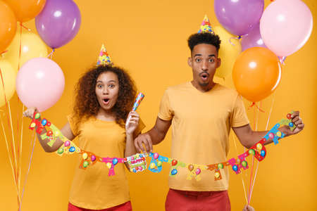 Shocked young friends couple african american guy girl in casual clothes birthday hat isolated on yellow background. Holiday party concept. Celebrating with colorful air balloons, pipe, paper garland.の写真素材
