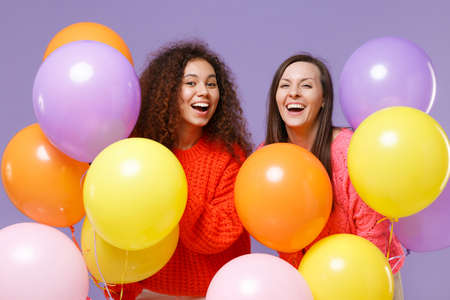 Cheerful two european african american women friends in knitted sweaters isolated on violet purple background. Birthday holiday party, people emotions concept. Celebrating hold colorful air balloons.の写真素材