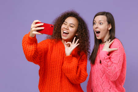 Excited funny young european african american women friends in knitted sweaters isolated on violet purple background. People lifestyle concept. Doing selfie shot on mobile phone, showing OK gesture.の写真素材