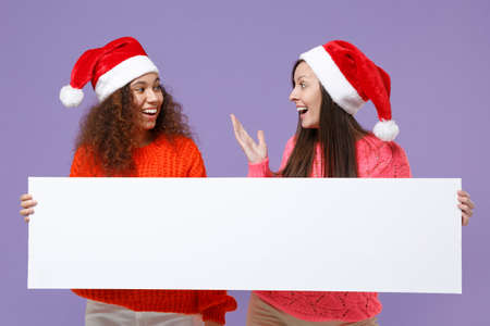 Excited european african american women friends in Christmas hat isolated on violet purple background. Happy New Year holiday concept. Mock up copy space. Hold blank sign board with place for text.の写真素材