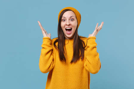 Amazed young woman girl in yellow sweater and hat posing isolated on blue background studio portrait. People sincere emotions lifestyle concept. Mock up copy space. Keeping mouth open spreading hands.の写真素材
