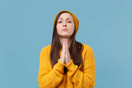 Pleading young woman girl in yellow sweater and hat posing isolated on blue background studio portrait. People sincere emotions lifestyle concept. Mock up copy space. Holding hands folded in prayer.の写真素材