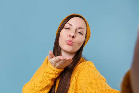 Close up of young woman in sweater and hat posing isolated on blue background studio portrait. People lifestyle concept. Mock up copy space. Doing selfie shot on mobile phone blowing sending air kiss.の写真素材