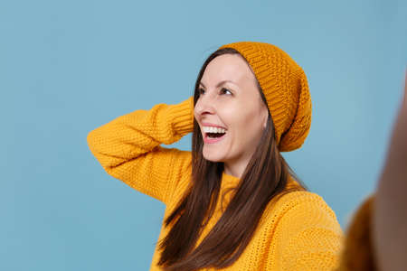 Close up of laughing young woman in sweater hat posing isolated on blue background studio portrait. People lifestyle concept. Mock up copy space. Doing selfie shot on mobile phone put hand on head.の写真素材
