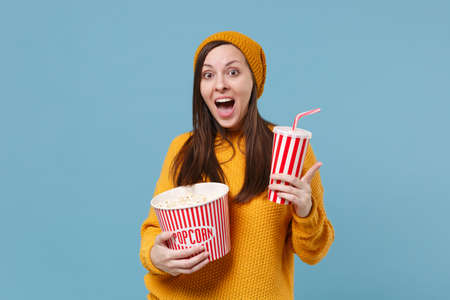 Excited young brunette woman in sweater hat posing isolated on blue background. People emotions in cinema lifestyle concept. Mock up copy space. Watching movie film hold bucket of popcorn cup of soda.の写真素材