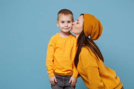 Woman in yellow clothes kissing kiss cheek cute child baby boy 4-5 years old. Mommy little kid son isolated on blue background studio portrait. Mothers Day love family parenthood childhood concept.の写真素材