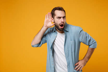 Shocked curious young bearded man in casual blue shirt posing isolated on yellow orange background, studio portrait. People lifestyle concept. Mock up copy space. Try to hear you with hand near ear.の写真素材