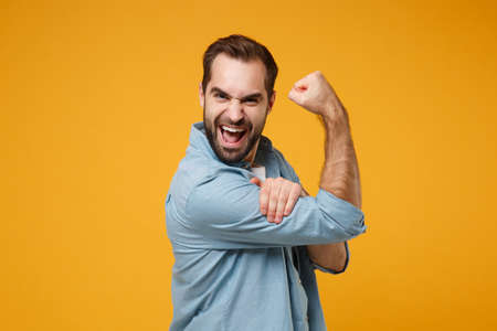 Cheerful young bearded man in casual blue shirt posing isolated on yellow orange wall background studio portrait. People sincere emotions lifestyle concept. Mock up copy space. Showing biceps muscles.の写真素材