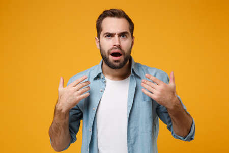 Amazed young bearded man in casual blue shirt posing isolated on yellow orange background, studio portrait. People emotions lifestyle concept. Mock up copy space. Keeping mouth open, spreading hands.の写真素材